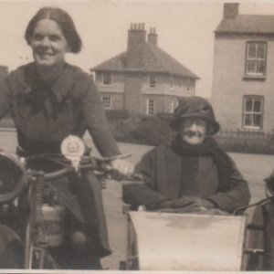This photograph was taken at the entrance to Church Lane.
It shows Beatrice Wright riding the motorcycle with her mother in law in the sidecar.
Beatrice ran the fish and chip shop, which was originally at that end of the village, with her husband Alf.
The photograph was probably taken some time in the 1920s.