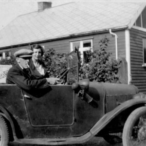 This photograph shows Henry Marshall, who owned the corner shop on Sea Dyke Way and Littlefeld Lane, sitting in his car outside the blue bungalow which used to stand in Littlefield Lane, to the rear of his shop.  A house now stands on this site.  To the right of the photograph the lady with the baby is Beattie, Henrys first wife, who sadly died as a result of a fire. Their son, Charles survived.  
The photograph was thought to have been taken around 1934. Also in the background, behind Beattie, can be seen a horse drawn vehicle which was used in his business.